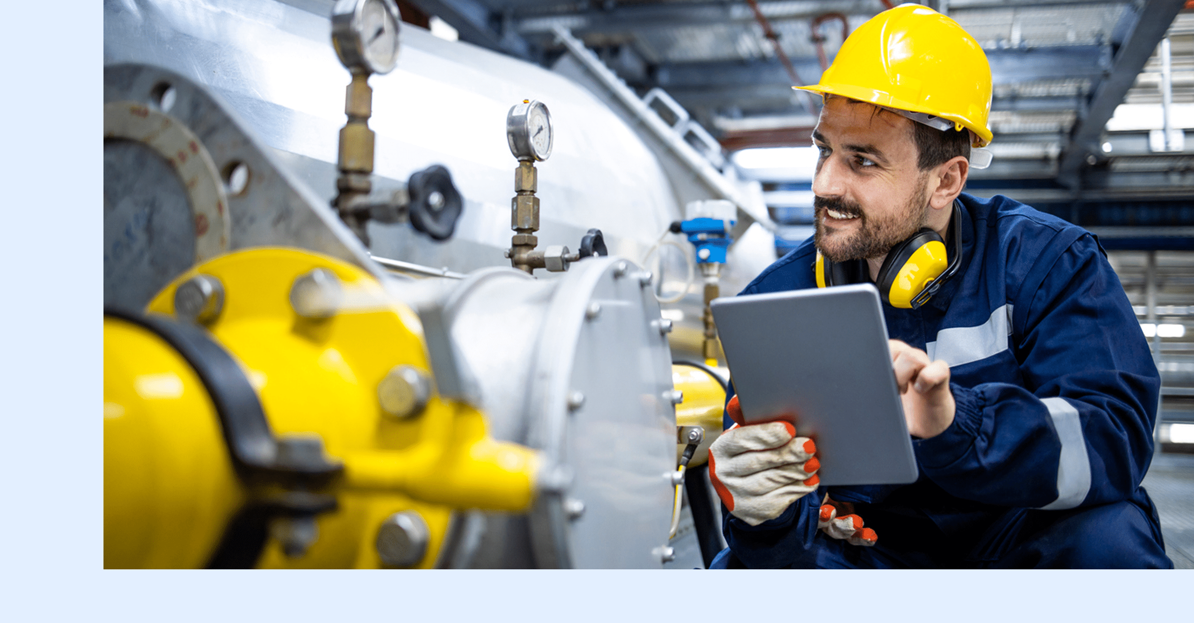 A worker with a hard hat, headphones, and gloves checks a tablet beside industrial equipment.