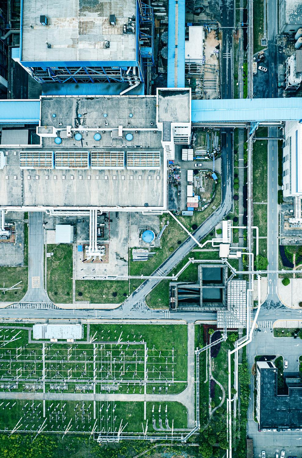 Aerial view of an industrial facility with large buildings, rooftop equipment, roads, green grassy areas, and a network of white pipelines running throughout the site.
