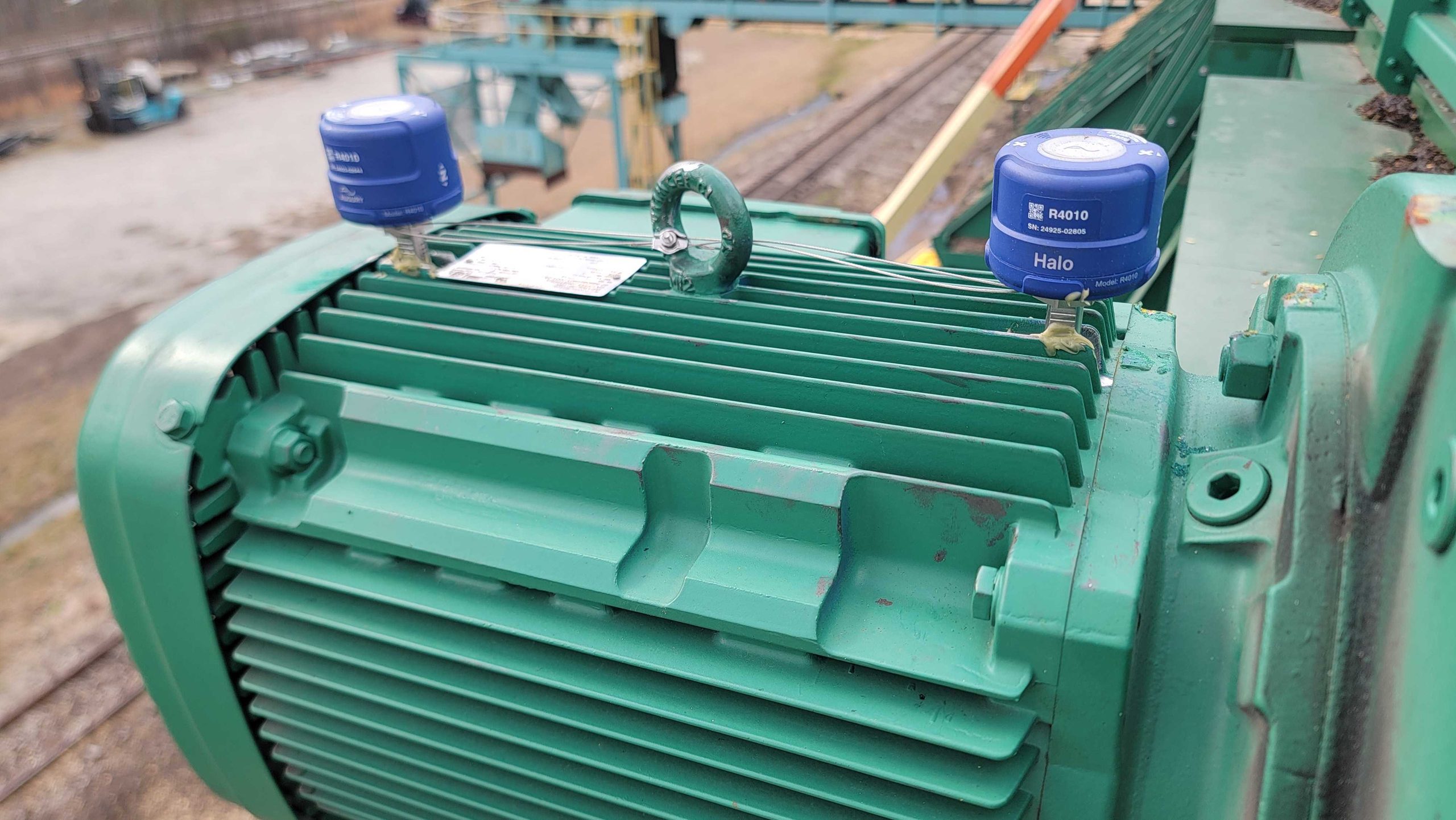 Close-up of a large green industrial electric motor with two blue caps labeled RA010 Halo on top, positioned outdoors near train tracks and construction equipment.