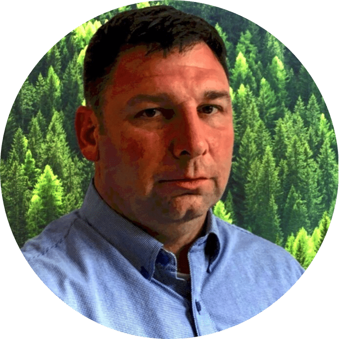 A man with short dark hair and a serious expression is wearing a light blue collared shirt. The background features dense green pine trees, emphasizing the importance of preventative maintenance to eliminate unplanned downtime.