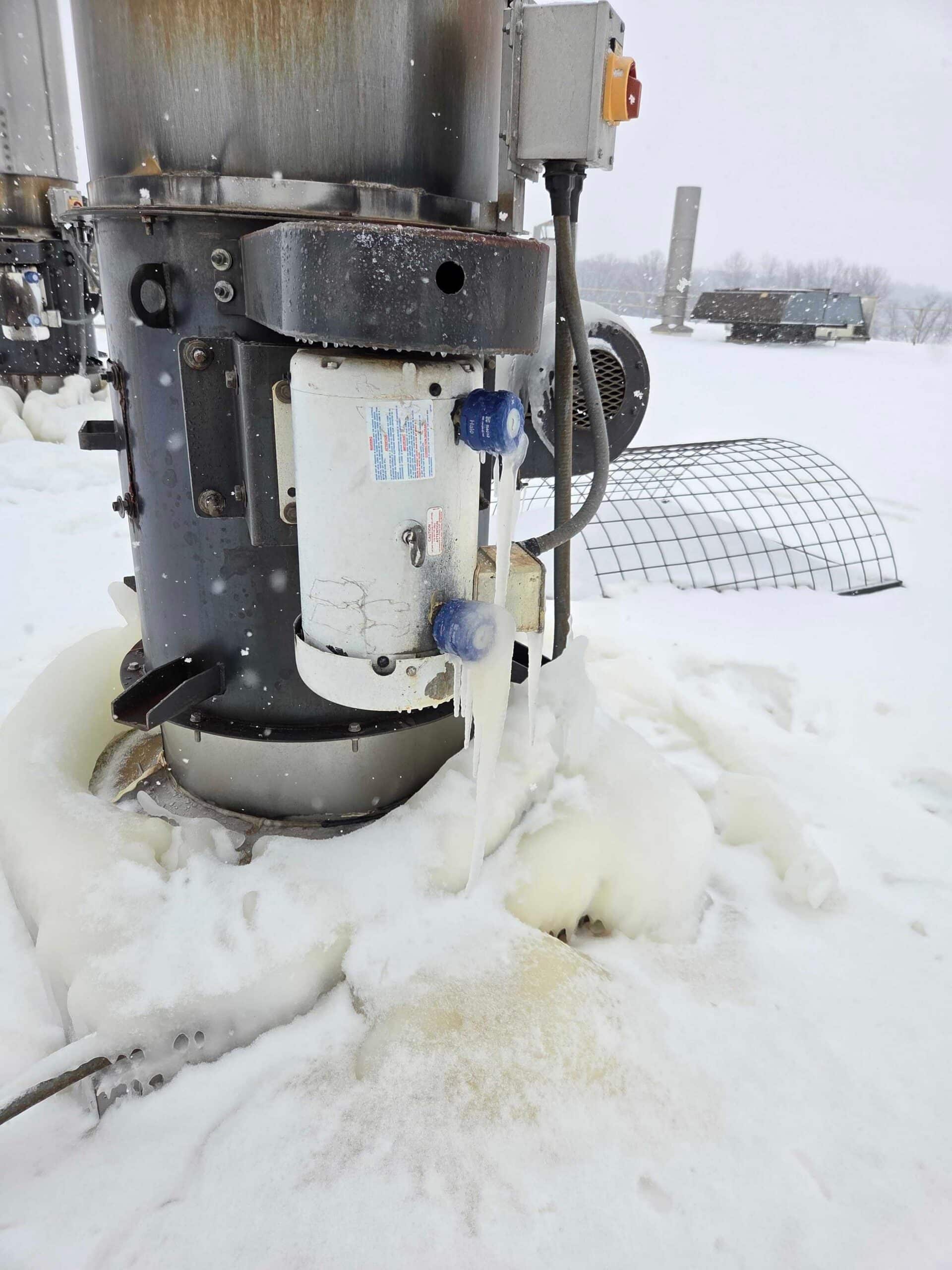 A close-up of industrial equipment outdoors, covered in snow and ice during winter. Icicles hang from tubes attached to the machinery, and snow blankets the ground and surrounding equipment.