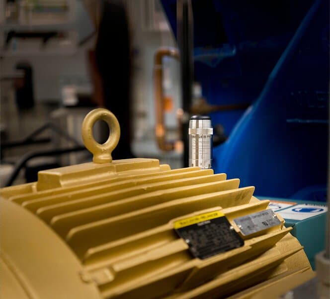 A close-up of a yellow industrial machine with cooling fins and attached labels; a small metallic cylindrical part is resting on top, with blurred equipment in the background.