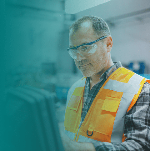 A man wearing safety glasses and an orange reflective vest works at a computer or control panel in an industrial or workshop setting.
