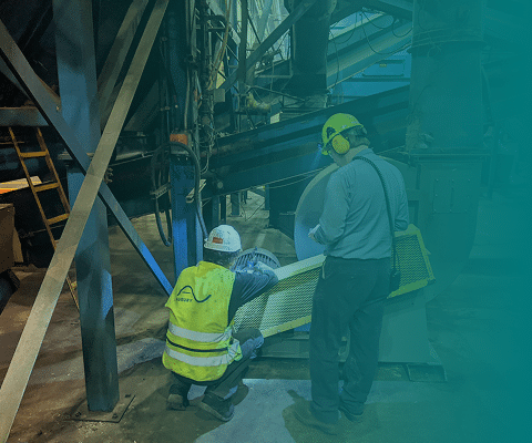 Two workers wearing safety gear inspect machinery in an industrial setting. One is squatting and the other stands beside him; both are observing the equipment closely under dim lighting.