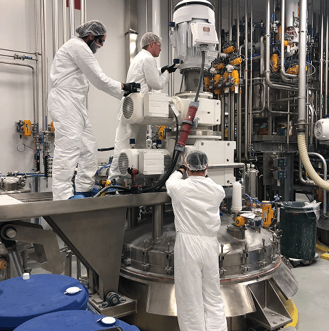 Three people in white protective suits and hairnets work on industrial equipment in a facility with many pipes and machinery. They appear to be inspecting or maintaining a large stainless steel vessel.
