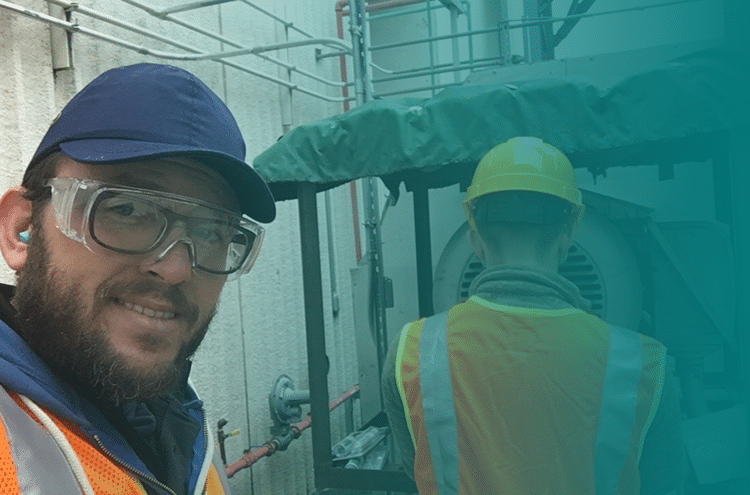 Two workers in safety gear stand near industrial equipment. One faces the camera smiling, wearing a blue cap, goggles, and orange vest; the other faces away, wearing a yellow hard hat and reflective vest.
