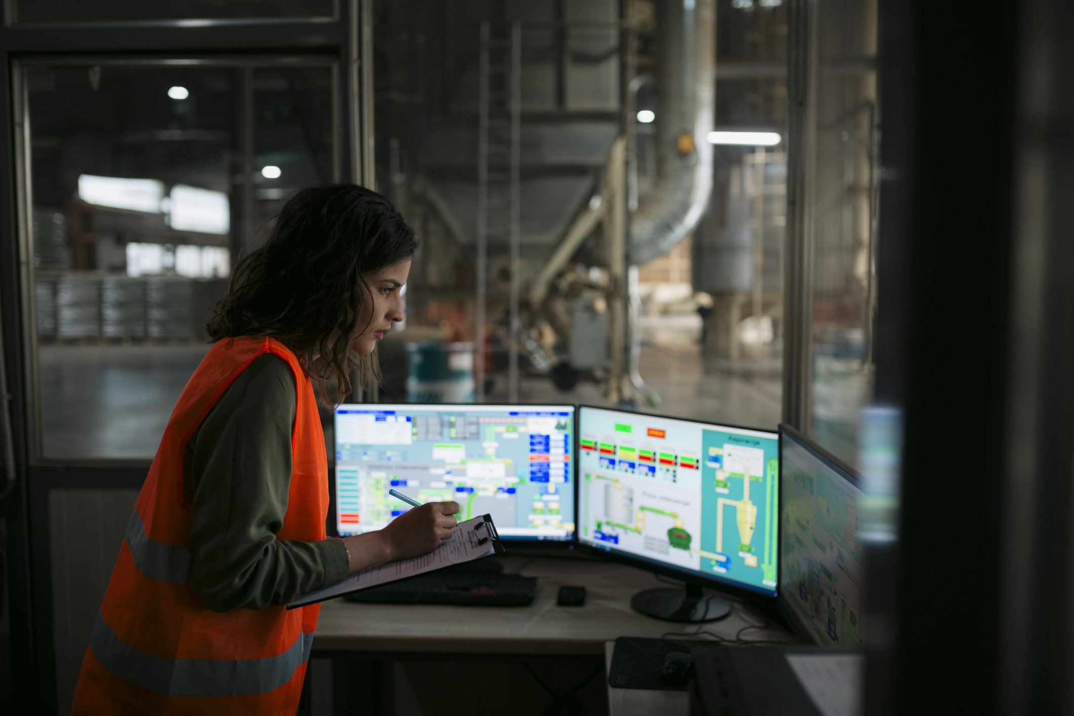 A person in an orange safety vest reviews information on multiple monitors and writes on a clipboard inside an industrial control room with a large factory visible through the window.