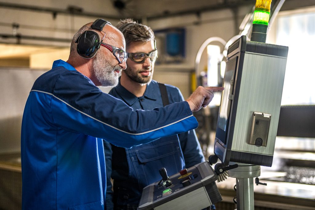 Two men in work uniforms and safety glasses operate a touchscreen control panel in an industrial setting, with one man pointing at the screen while the other observes closely.