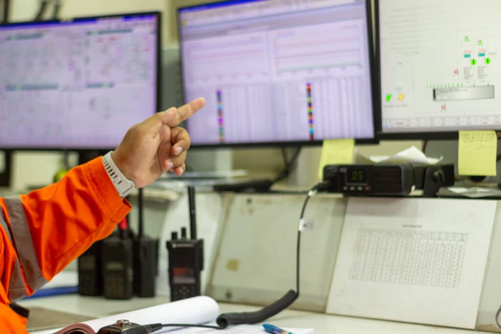 A person in an orange safety jacket points at data displayed on multiple computer monitors in a control room, with charts, graphs, and equipment visible on the desk.