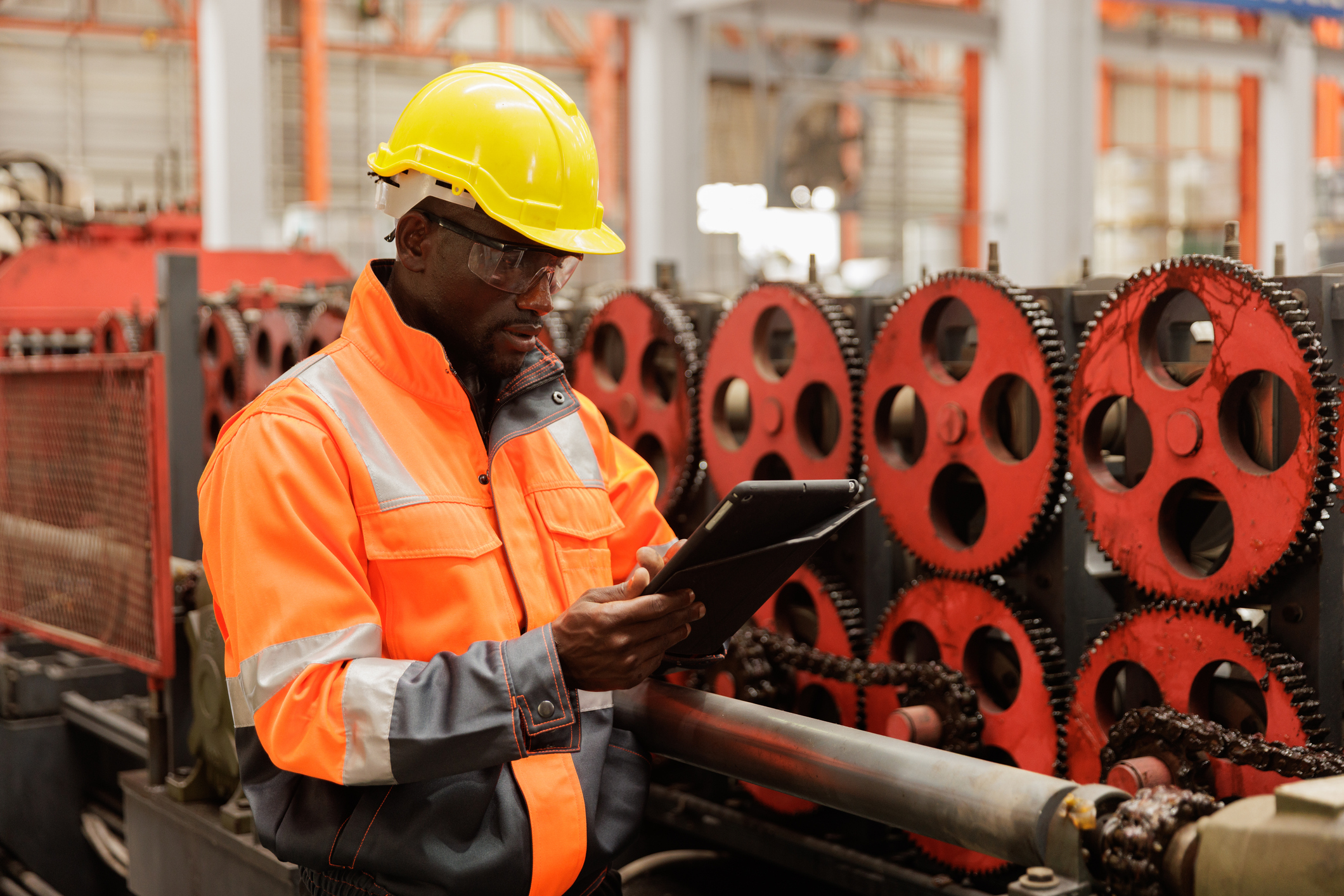 A worker in an orange safety jacket and yellow helmet inspects a tablet in a factory, standing next to red industrial gears and machinery.