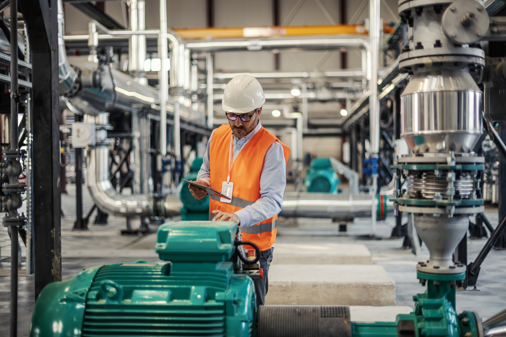 A man in a hard hat and orange vest uses a tablet while inspecting industrial machinery and pipes in a large factory setting.