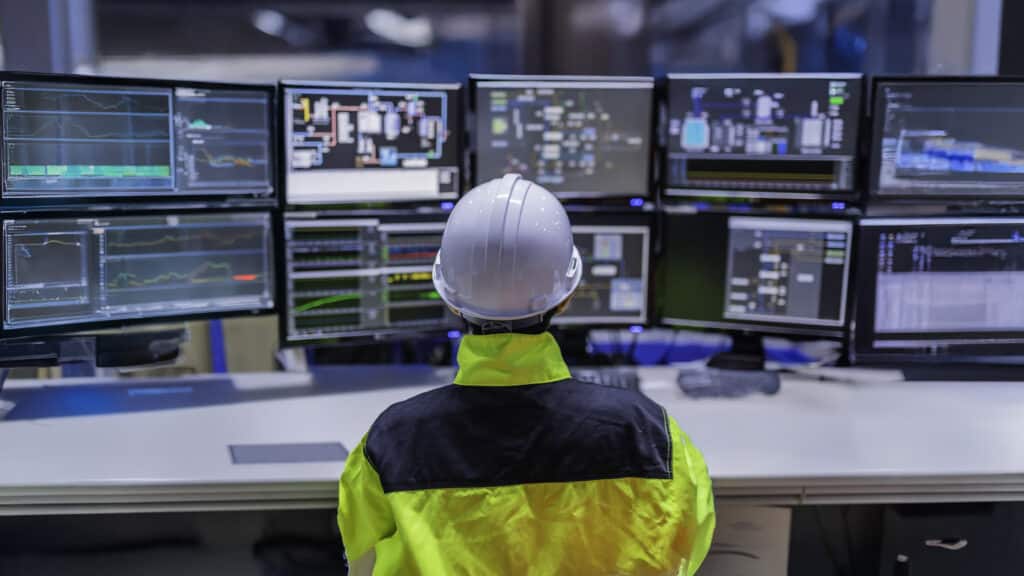 A person wearing a white hard hat and high-visibility jacket sits at a desk facing multiple large computer monitors displaying technical data and control panels in a control room.