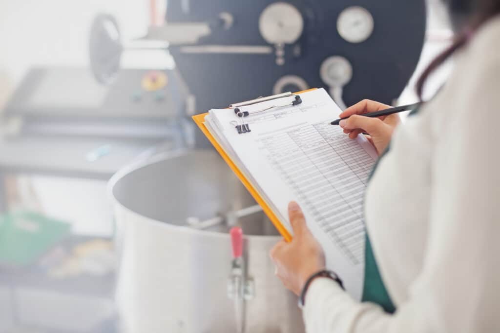 A person holding a clipboard and pen fills out a checklist or log sheet in front of industrial machinery with dials and gauges, possibly in a manufacturing or laboratory setting.
