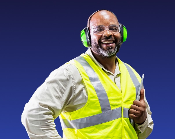A smiling man wearing a yellow reflective safety vest, protective glasses, and green earmuffs holds a tablet against a solid dark blue background.