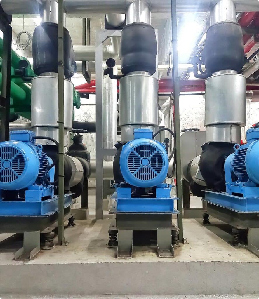 Three large blue industrial pumps are installed on a concrete platform, with silver pipes connected above them and various other pipes and machinery visible in the background.