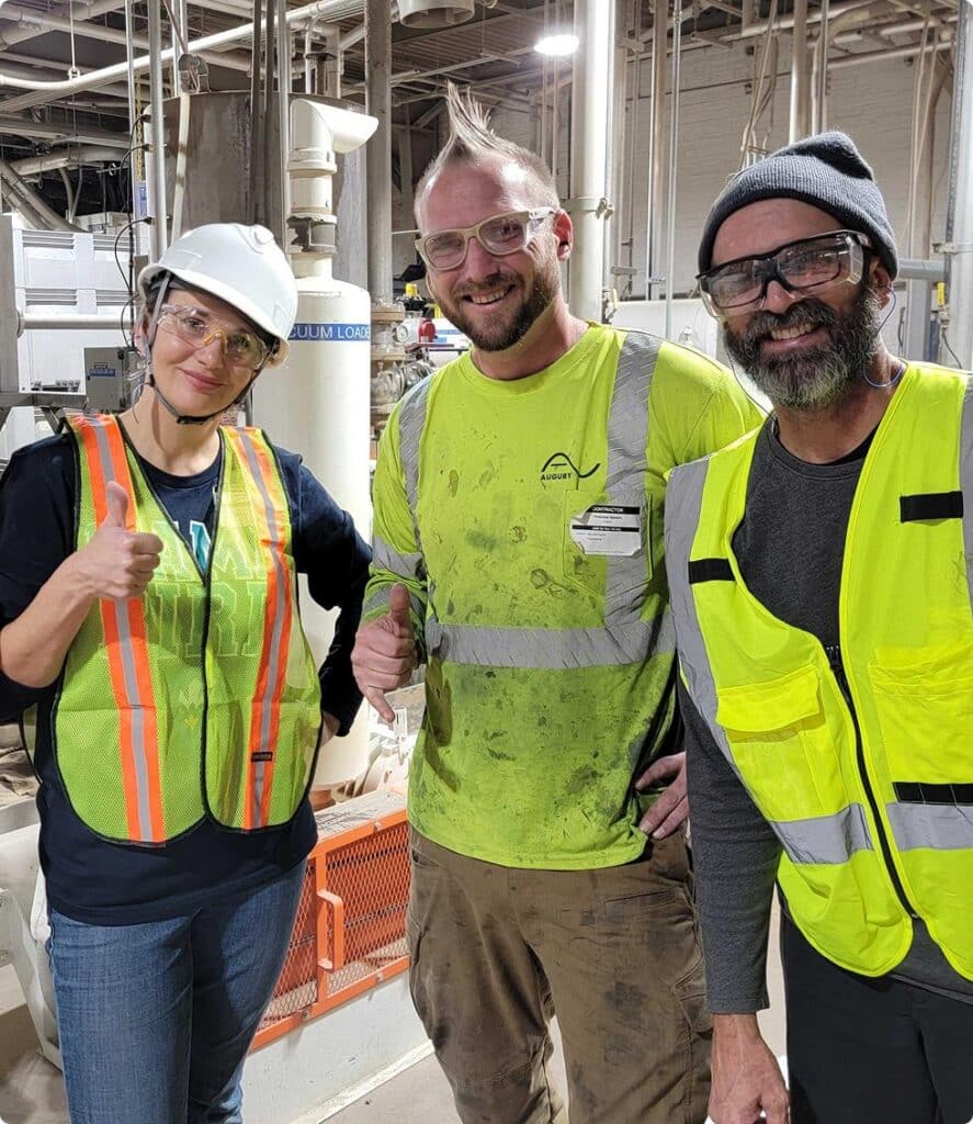 Three workers in safety gear, including hard hats, safety glasses, and reflective vests, smile and give thumbs up while standing together in an industrial setting with equipment in the background.