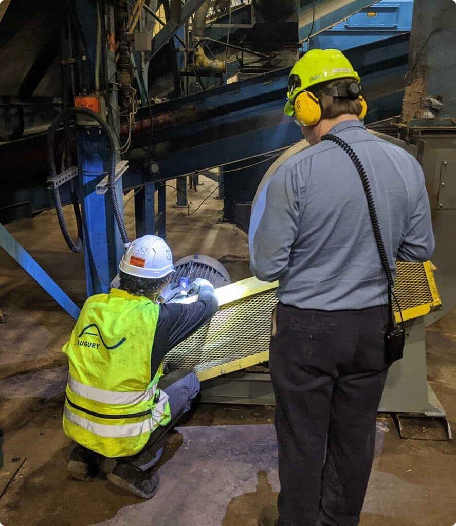 Two workers wearing safety helmets and high-visibility gear inspect or repair machinery in an industrial setting. One kneels and examines equipment, while the other stands nearby with ear protection.