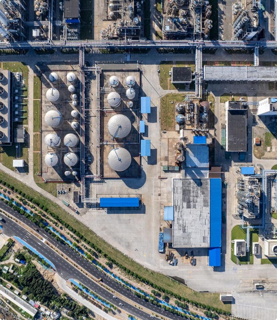 Aerial view of an industrial facility with large spherical tanks, pipes, buildings, and paved roads; green areas and trees surround the complex, with a highway visible at the bottom.