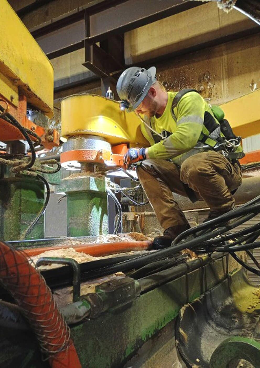 A construction worker in safety gear and a helmet kneels on machinery, inspecting or repairing equipment inside an industrial facility with metal beams and cables around him.