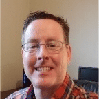 A man with glasses and a red plaid shirt is smiling in a well-lit room with a beige wall and a black office chair in the background, perhaps reflecting on improved reliability in machine health for the pulp and paper industry.