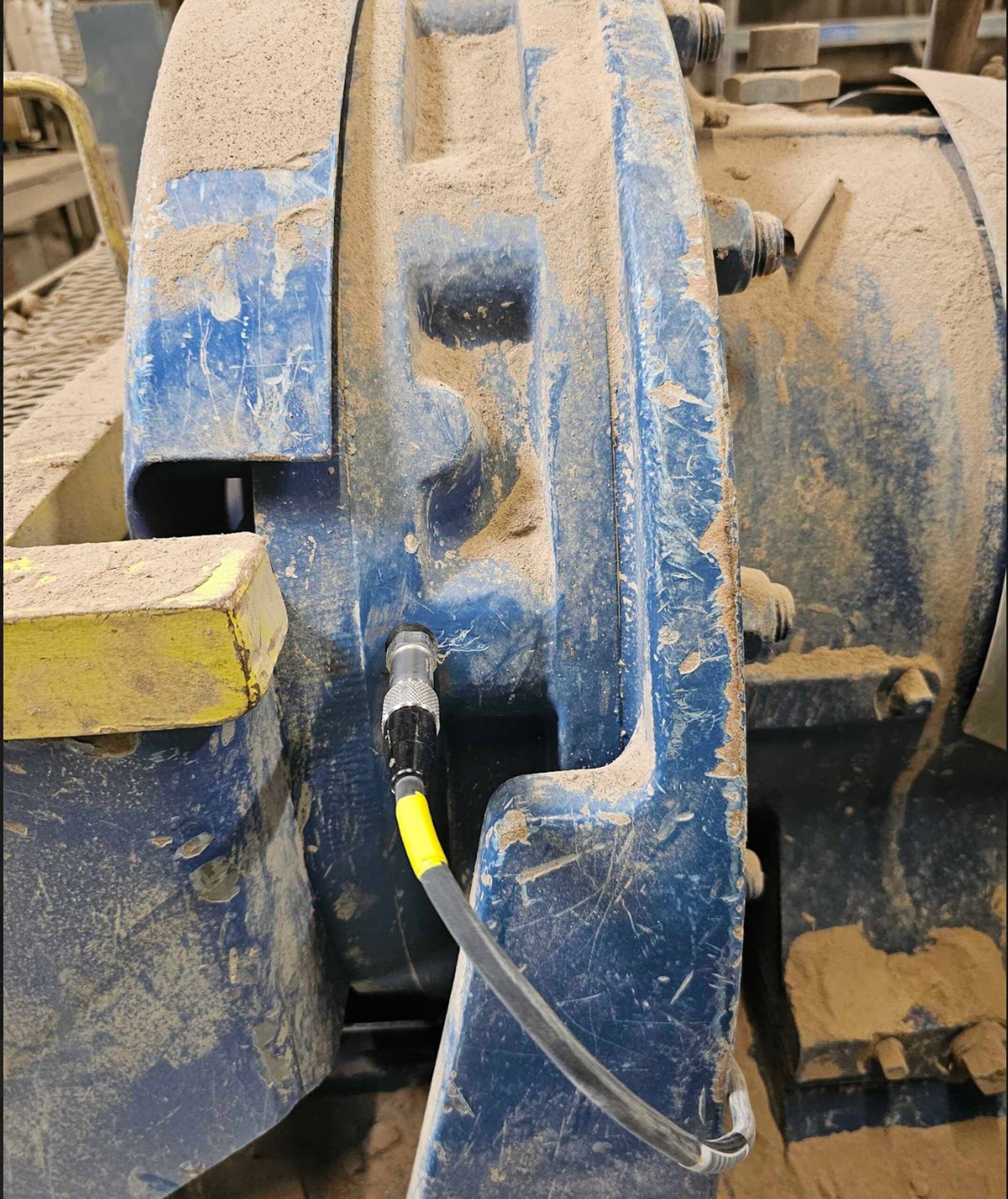 A close-up of a blue industrial machine covered in dust, showing a black cable with a yellow stripe connected to it. Large bolts and metal surfaces are visible, indicating a heavy-duty mechanical environment.