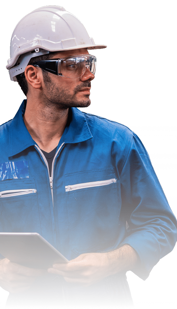 A man wearing a white hard hat, clear safety glasses, and a blue coverall holds a tablet and looks to the side, appearing focused.
