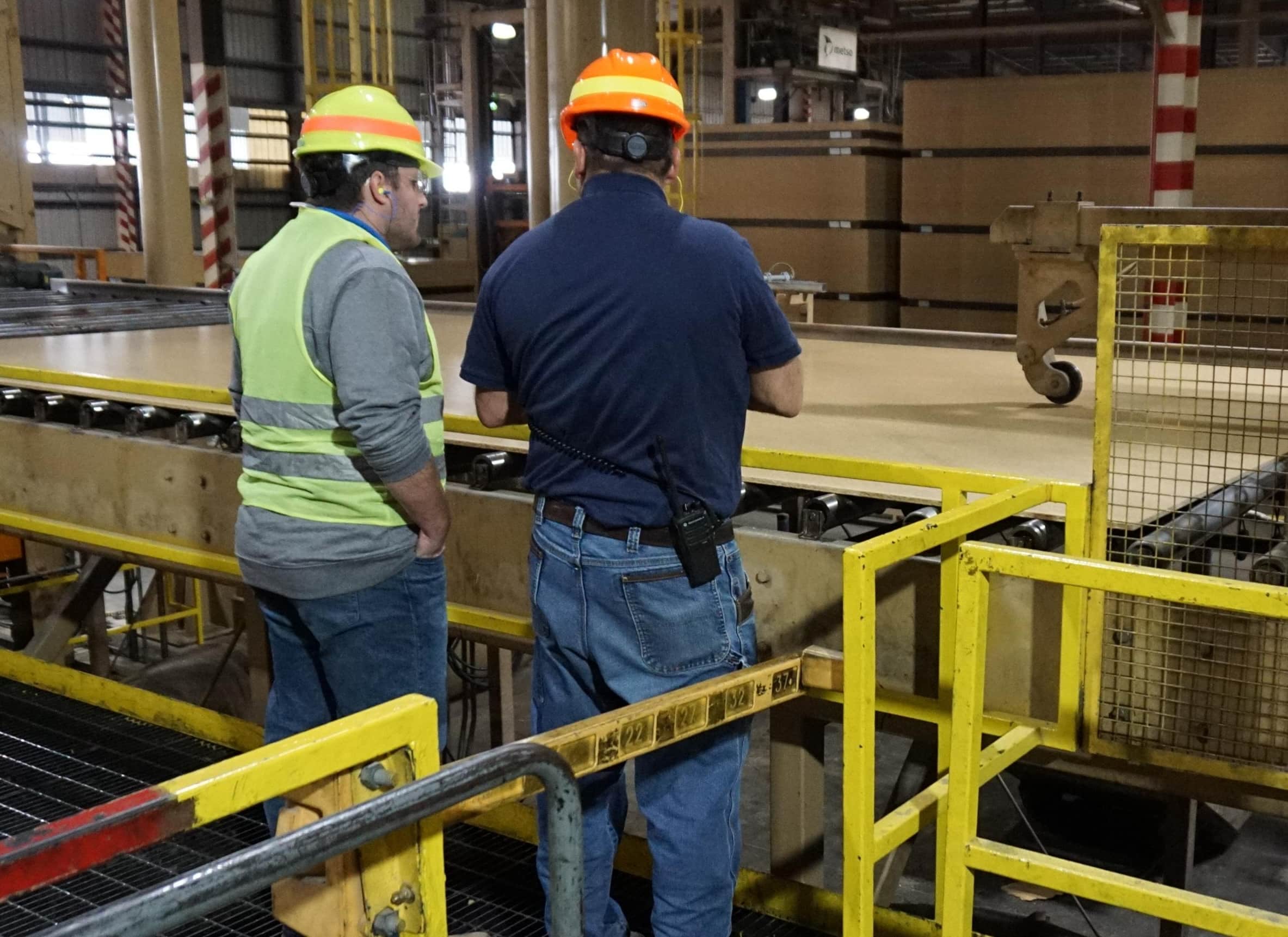 Two workers wearing safety equipment, including helmets and vests, stand in front of a conveyor belt in a factory or industrial setting, observing a large sheet of material being processed.